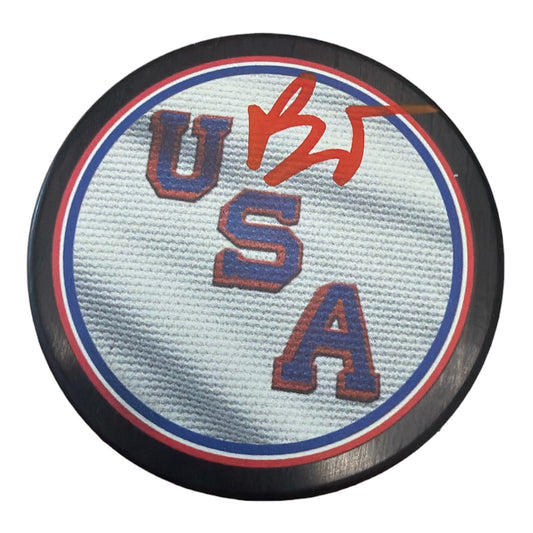 A circular USA hockey puck with a white textured center, blue and red borders, and red embroidered letters spelling USA, with a red autograph on top, shown on a white background.