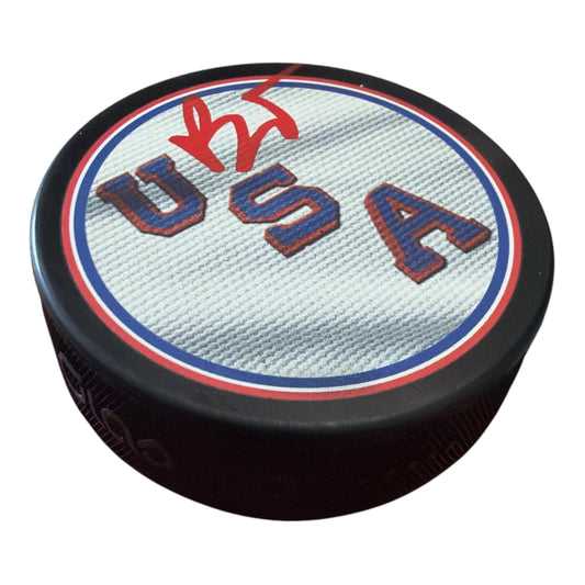 A black hockey puck featuring a printed USA flag design on the top, with 'USA' in red and blue outlined letters on a white textured background.